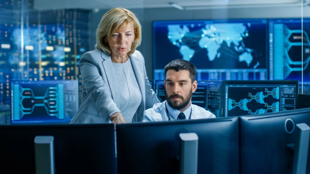 In the System Monitoring Room, a cybersecurity product manager controls work of the Operator. They're Surrounded by Monitors Showing Relevant Technical Data.