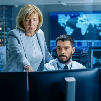 In the System Monitoring Room, a cybersecurity product manager controls work of the Operator. They're Surrounded by Monitors Showing Relevant Technical Data.